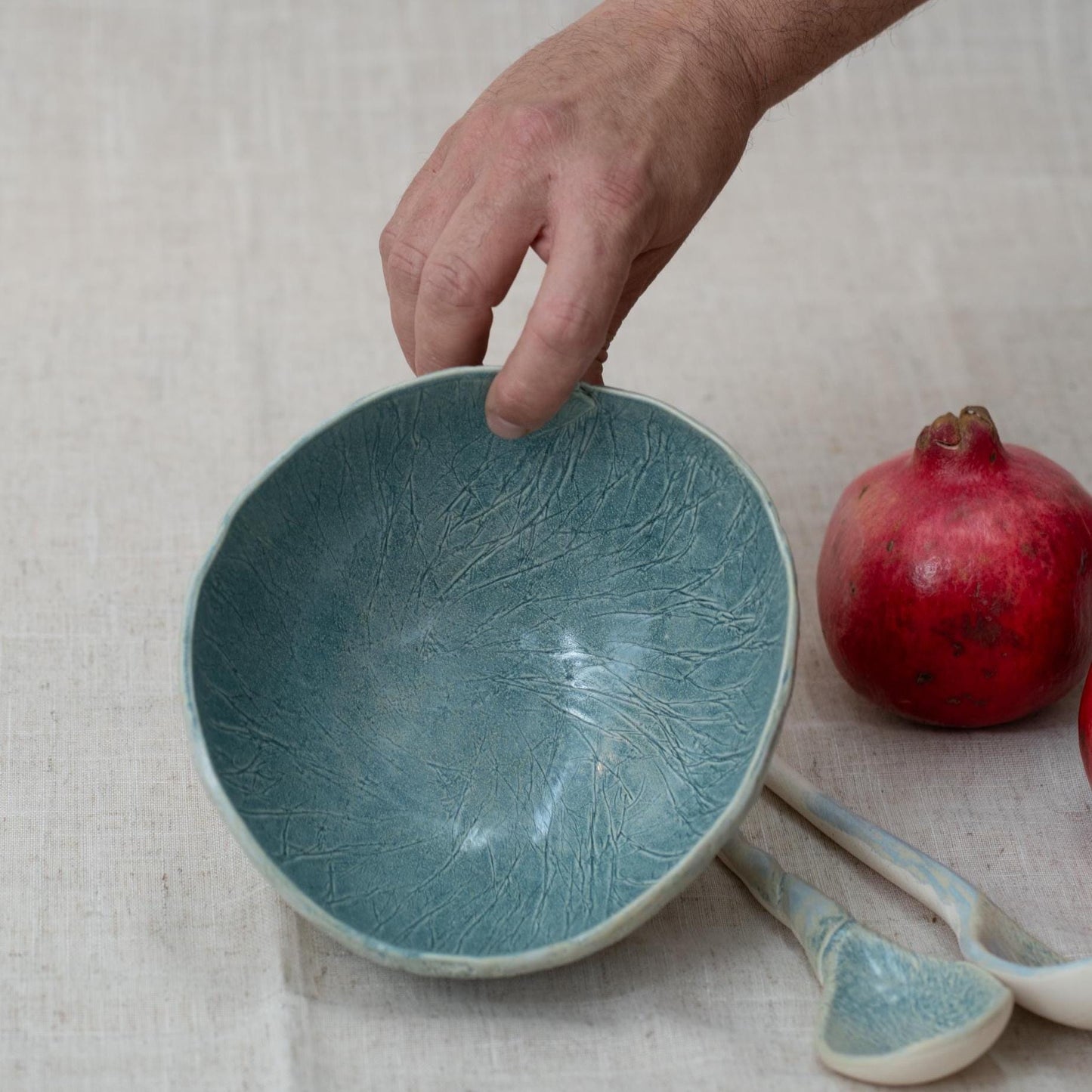 Hand holding a textured blue ceramic bowl with a pomegranate and spoon on a light fabric background