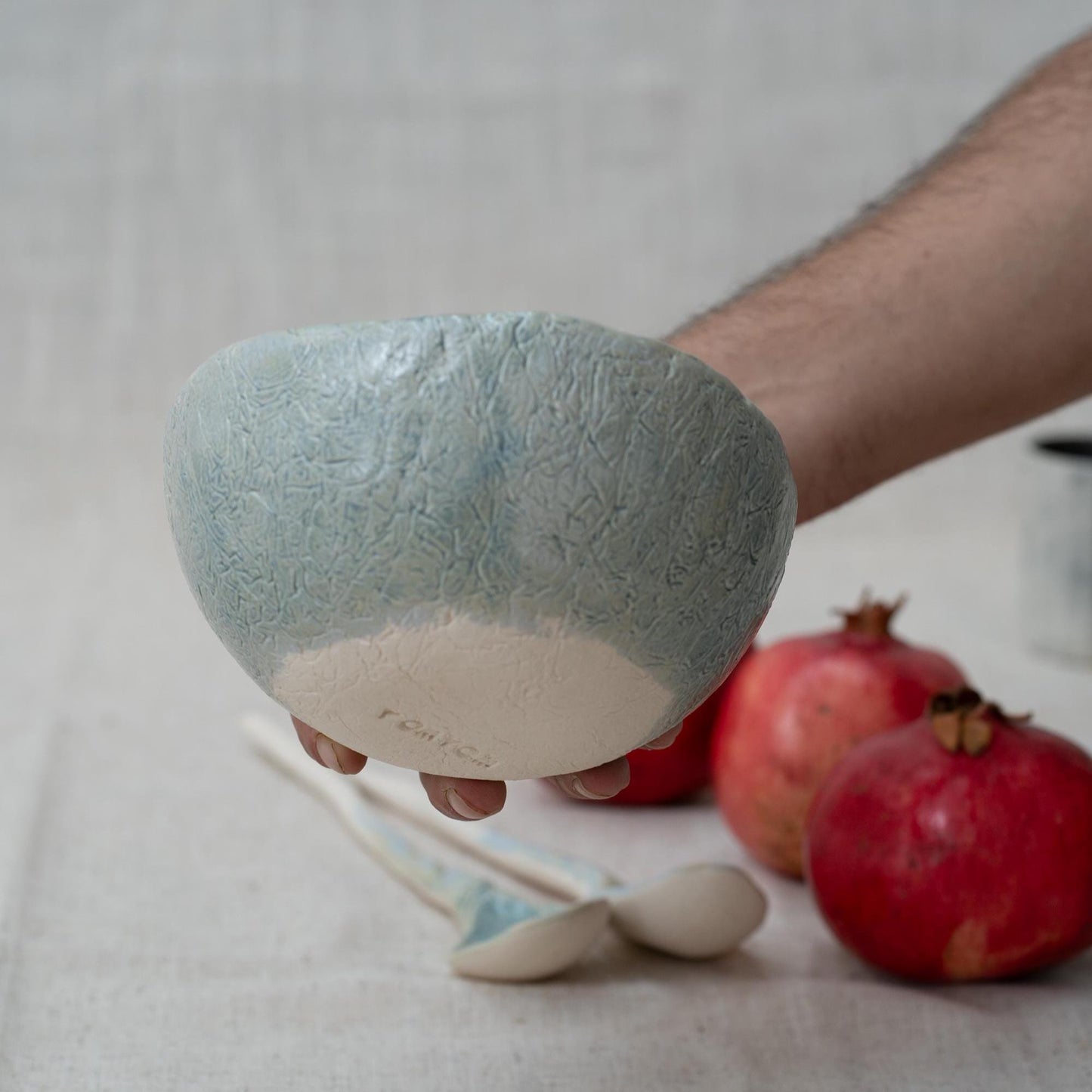 Person holding a textured ceramic bowl with pomegranates and spoons on a neutral background