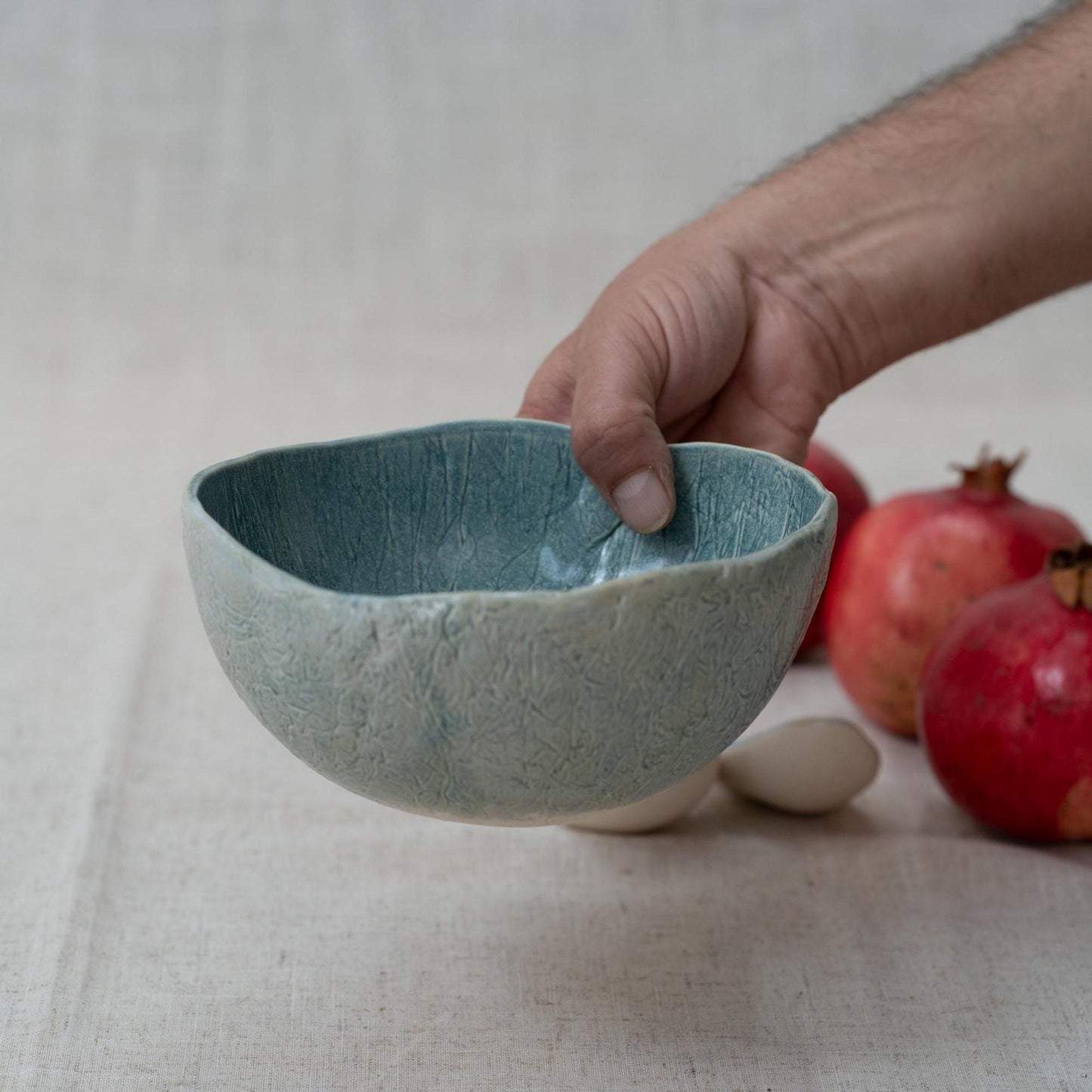 Hand holding a textured ceramic bowl with pomegranates on a neutral background