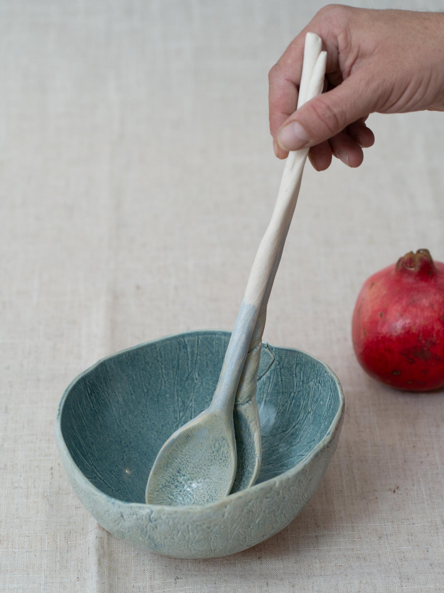 Ceramic bowl with two ceramic spoons inside, held by a hand, on a textured surface with a pomegranate.