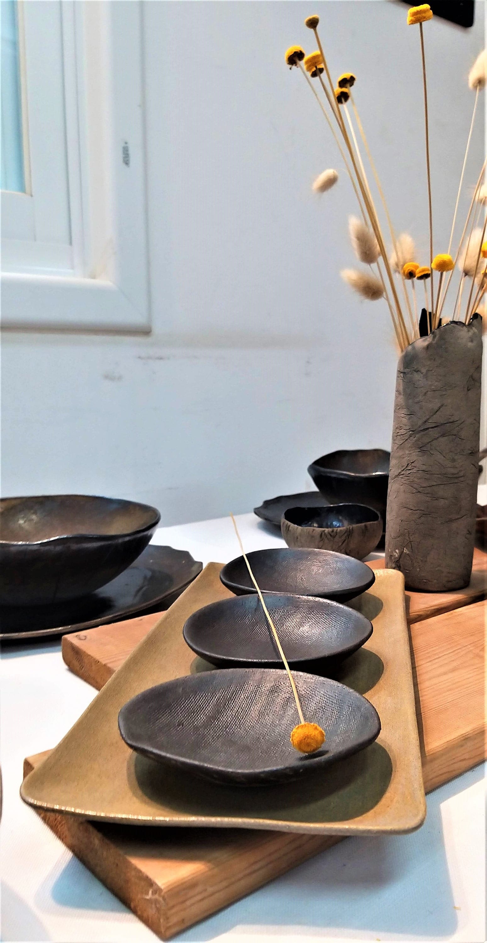 A mocha brown rectangular ceramic tray on which are placed three black ceramic bowls next to a vase and two black ceramic plates on each of which is placed a black ceramic soup bowl