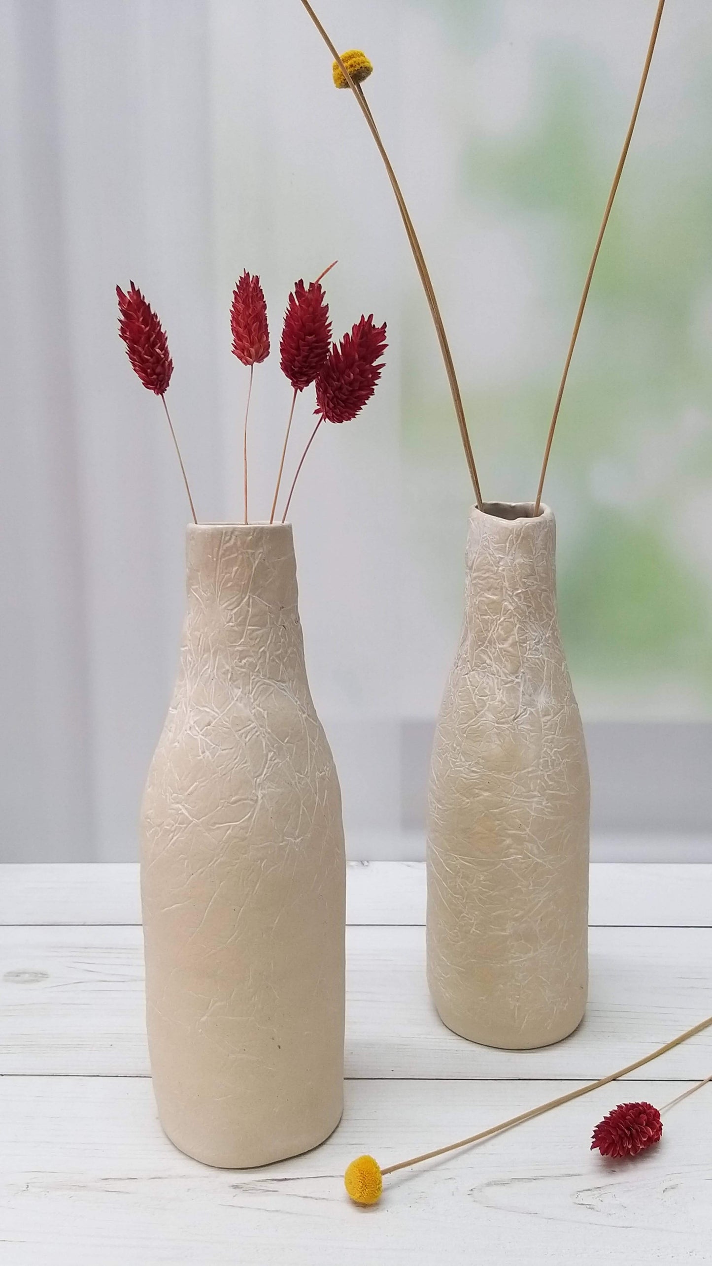 Two bottle-shaped ceramic white vases are placed next to each other with red and yellow flowers inside